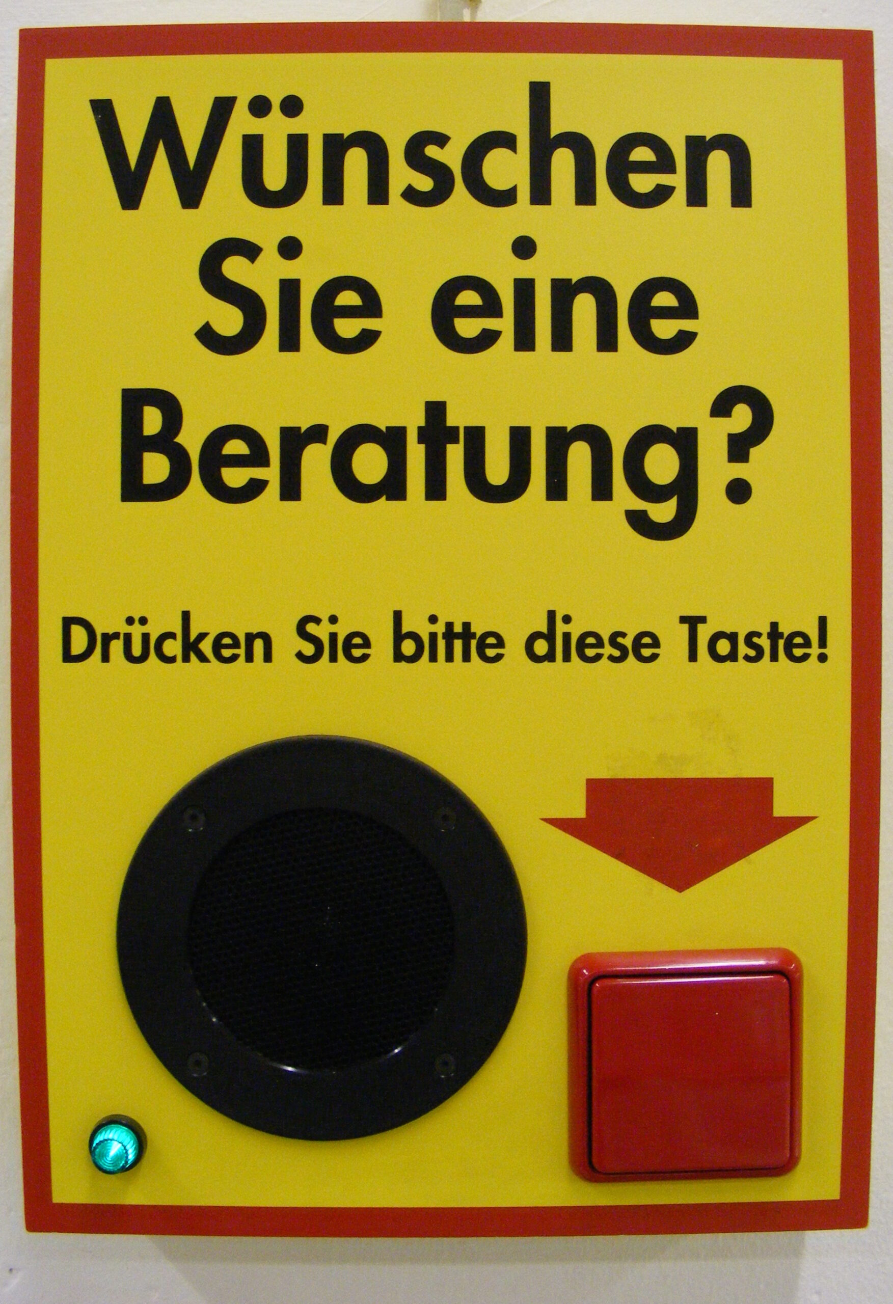 Ein Schild mit der Aufschrift "Wünschen Sie eine Beratung? Drücken Sie bitte diese Taste!" In das Schild ist ein kleiner Lautsprecher integriert und eine rote Taste, auf die ein ebenfalls roter Pfeil zeigt.