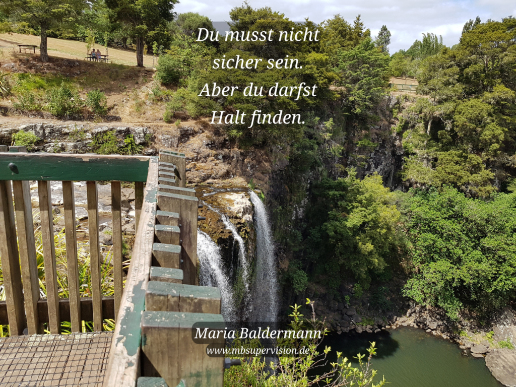 Eine Landschaft mit Wasserfall von oben. Links m Vordergrund ist die Brüstung der Aussichtsplattform zu sehen. Dazu der Text: Du musst nicht sicher sein. Aber du darfst Halt finden.