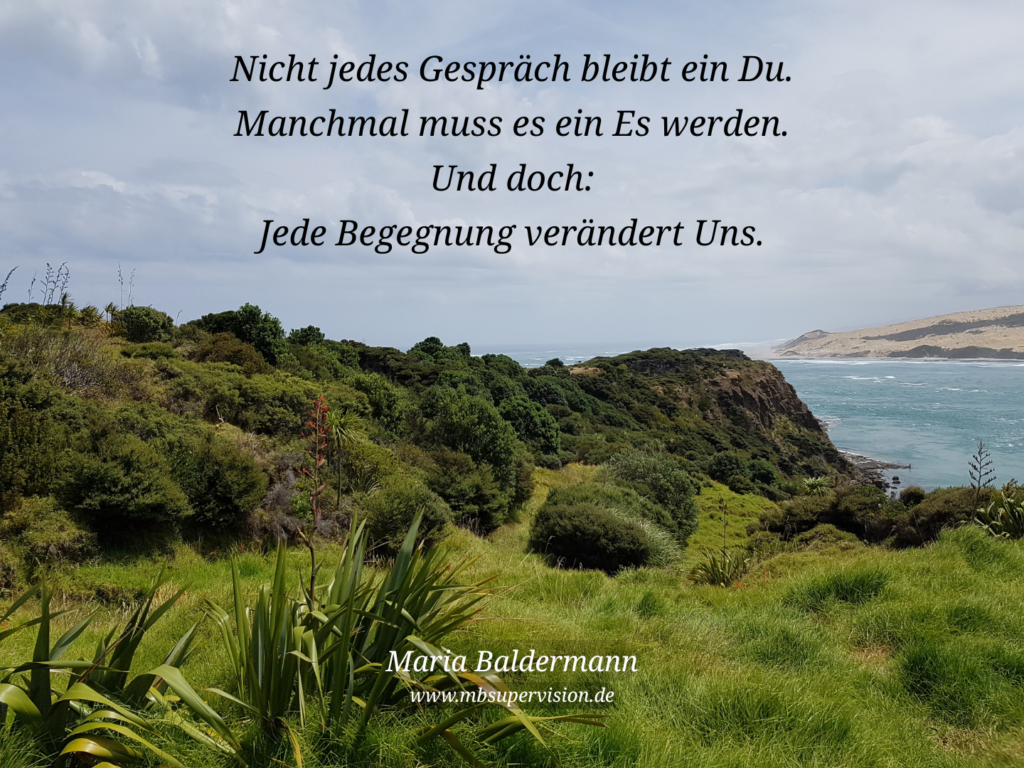 Eine hügelige Landschaft mit Gras und Büschen, dahinter ein Meeresarm. Davor der Text: Nicht jedes Gespräch bleibt ein Du. Manchmal muss es ein Es werden. Und doch: Jede Begegnung verändert Uns.