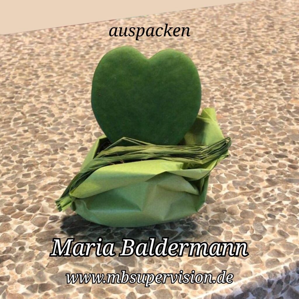 Ein grünes, herzförmiges Objekt steckt in einer gefalteten Papierhülle, die auf einer gemusterten Oberfläche liegt. Oberhalb steht das Wort 'auspacken'. Im unteren Bereich sind der Name 'Maria Baldermann' und die Internetadresse 'www.mbsupervision.de' zu lesen.