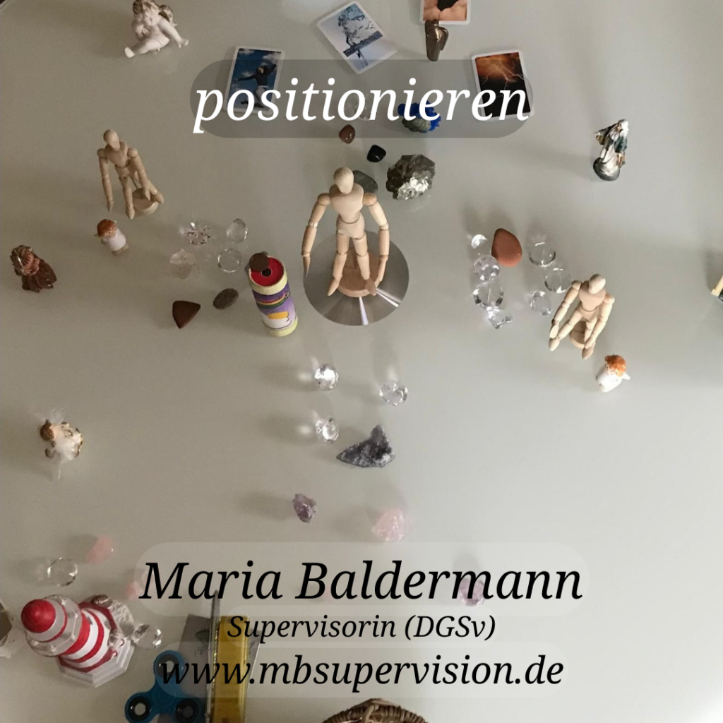 Auf einer hellen Tischfläche stehen mehrere kleine Holzfiguren, Steine, Glassteine und weitere Miniaturgegenstände verteilt. Zwei größere Holzmodelle in neutraler Pose befinden sich zentral. Im oberen Bereich steht das Wort 'positionieren'. Im unteren Bereich sind der Name 'Maria Baldermann', die Berufsbezeichnung 'Supervisorin (DGSv)' und die Webadresse 'www.mbsupervision.de' angegeben.